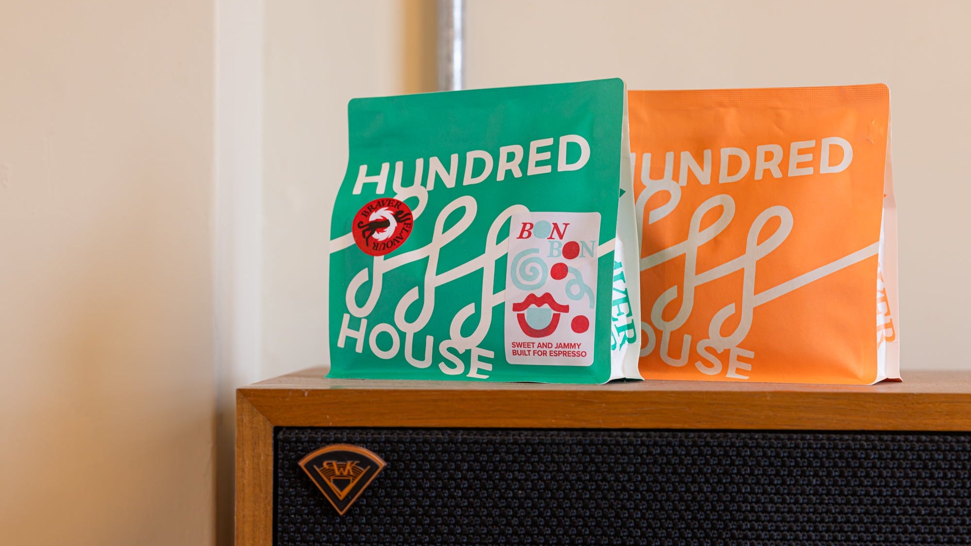Two 'Hundred House' coffee bags, one green and one orange, on a wooden surface.